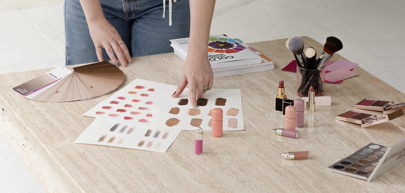 Melissa swatching foundation and lip shades onto paper beside a Pantone fan, brushes, lipsticks and eyeshadow palettes